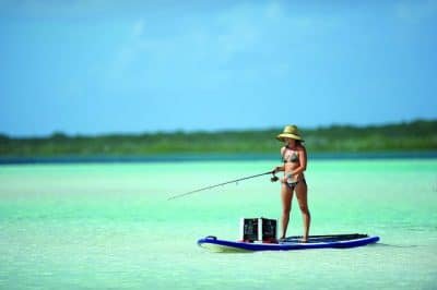 Young woman in a bikini and a straw hat fishes off of an SUP in shallow, tropical green waters