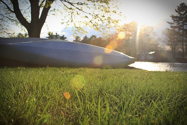 Large kayak laying on its side on the grass by a lake with a ray of sunlight hitting it as the sun sets. 