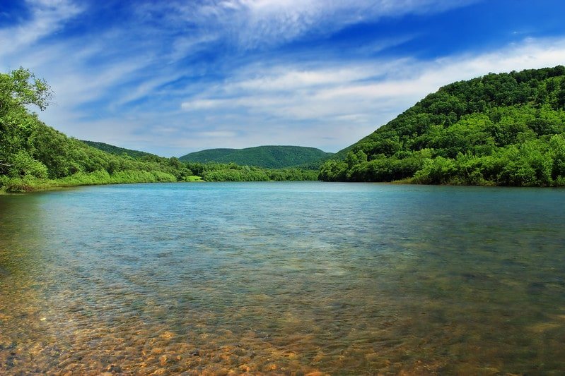 Lush green trees on hilly landscapes line the shallow, rocky bottom shores of the Susquehanna River. 