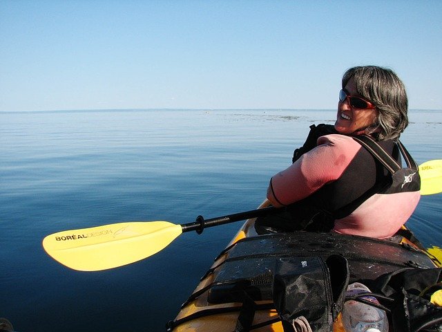 Older woman looks back for a photo as she kayaks a large, glassy lake